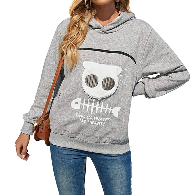 A woman wearing a gray hoodie with a cat face design and a built-in pouch for holding small pets, paired with blue jeans.