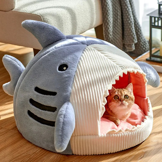 A plush gray cat bed shaped like a shark with an orange and white cat peeking out from the shark's mouth.