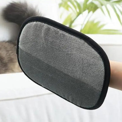 A hand holding a double-sided pet grooming glove with a mesh of bristles on a black background. A cat is visible in the background, and there is a plant in the top left corner.