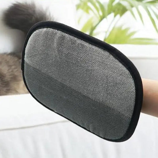A hand holding a double-sided pet grooming glove with a mesh of bristles on a black background. A cat is visible in the background, and there is a plant in the top left corner.