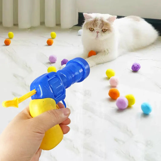 A hand holding a blue and yellow cat toy launcher with a yellow ball attached, in a room with a white cat lying down next to it.