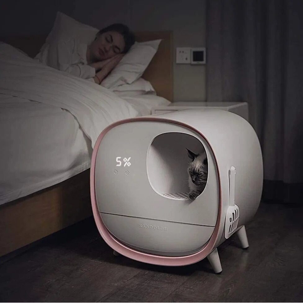 A modern white and pink self-cleaning pet litter box with an automatic cleaning feature, situated in a dark room next to a bed.