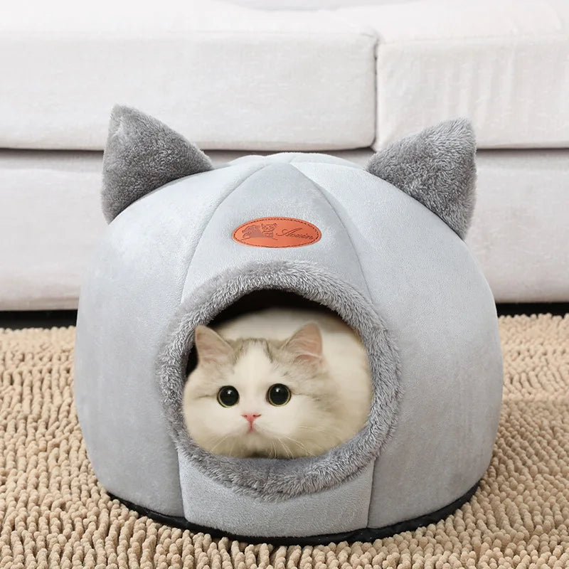 A white cat peeking out from a cozy, gray cat bed shaped like a house with a plush cushion inside.