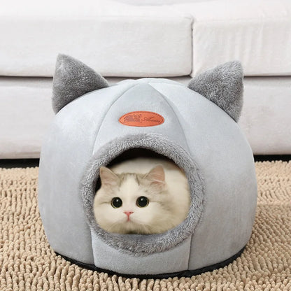 A white cat peeking out from a cozy, gray cat bed shaped like a house with a plush cushion inside.