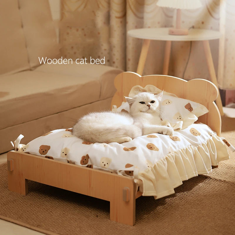 A white cat with a teddy bear design on the bedding, resting on a wooden cat bed with a brown and white blanket.